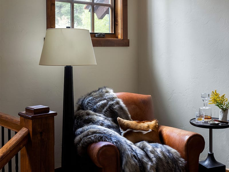 Nida Risto Interiors - Jackson Hole, Wyoming - Interior of a Snake River Sporting Club house showing a large leather armchair covered in a fury blanket set in a beam of light coming from a high window.